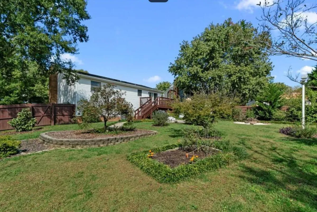 The image shows a peaceful, single-story house with a deck surrounded by flower beds, mature trees, and open yard space. The setting emphasizes the connection between the home and its natural surroundings, ideal for outdoor enjoyment.