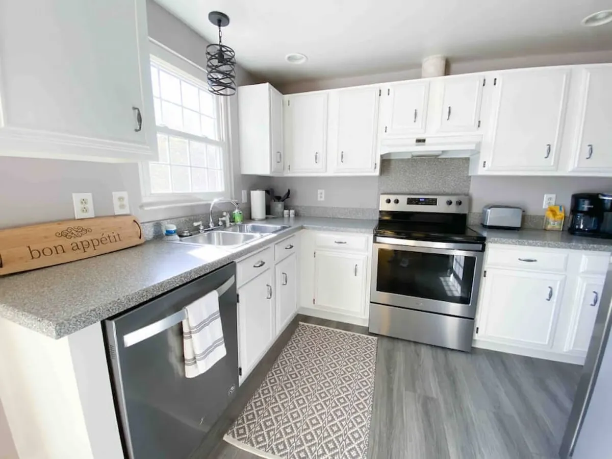 A stylish kitchen featuring stainless steel appliances, pristine white cabinetry, a decorative rug, and a 'bon appétit' sign. The scene is inviting, with ample counter space and hints at culinary activity, suggesting both gourmet cooking and casual family meals.