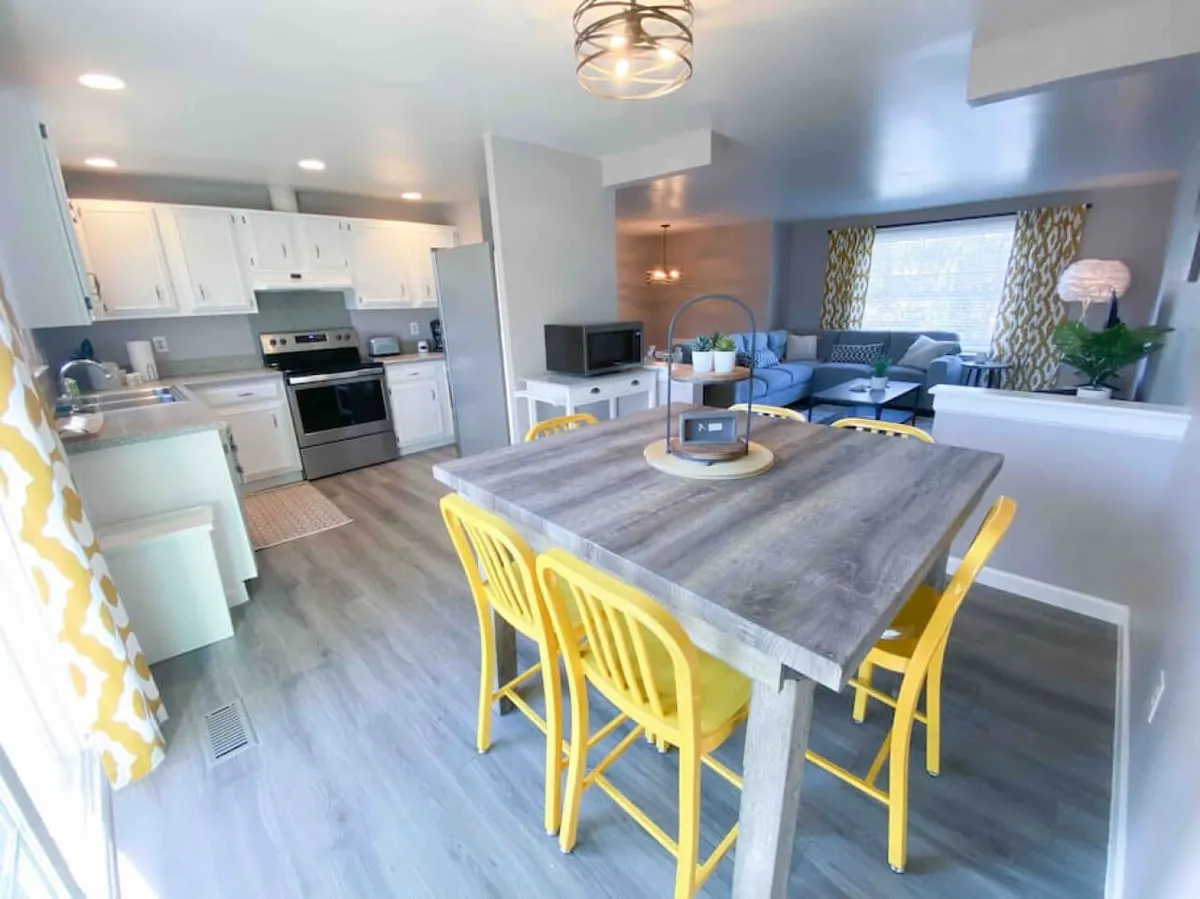 This image captures an open-plan living and dining area featuring a dining table with bright yellow chairs, a cozy living space with a blue couch, and modern kitchen appliances in the background. The overall scene is filled with natural light and exudes a cheerful, welcoming mood ideal for gatherings.