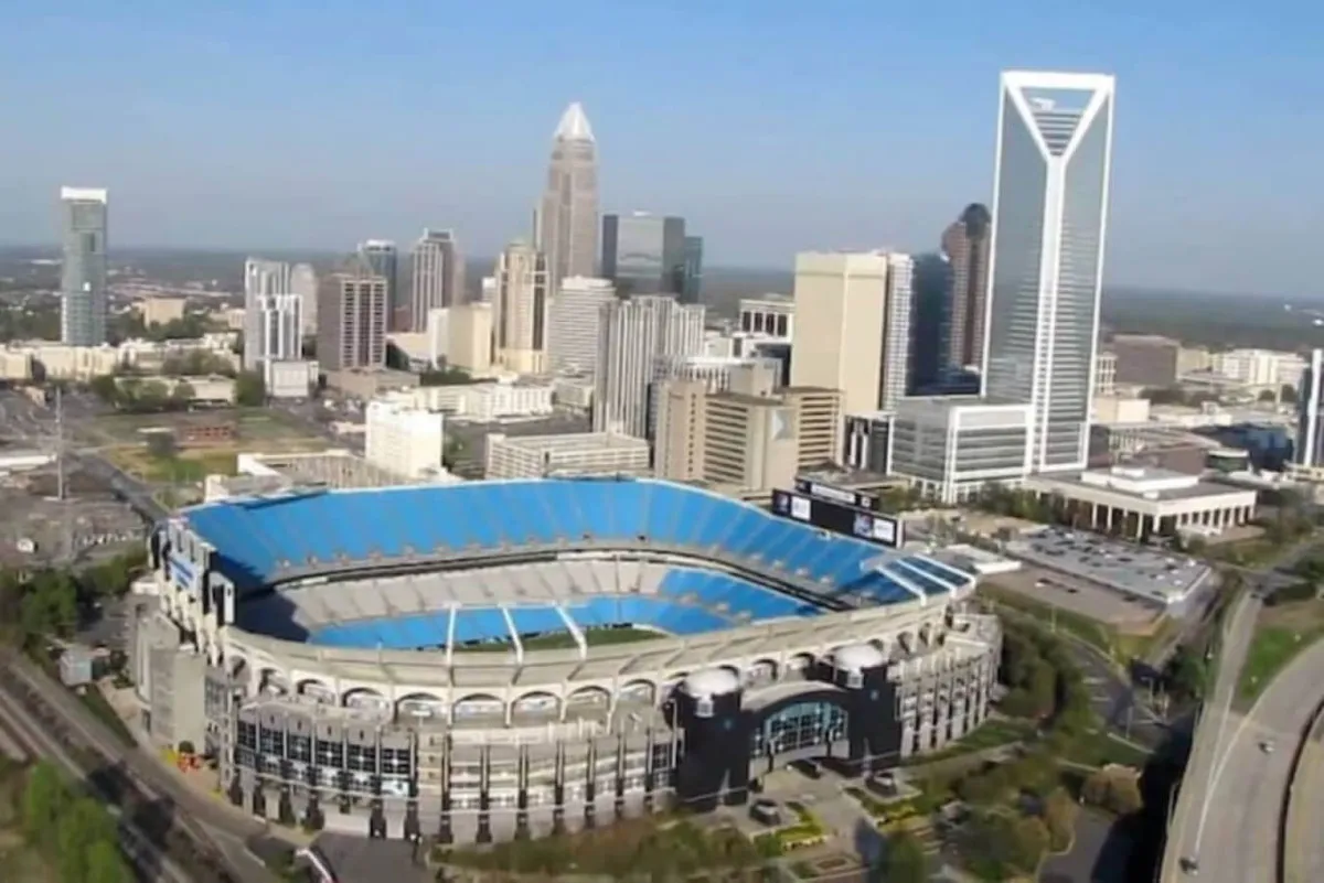 A panoramic view of the Charlotte skyline featuring the iconic Bank of America Stadium in the foreground and a cluster of high-rise buildings behind, capturing the city's dynamic energy. The image conveys a sense of excitement, urban vibrancy, and proximity to major attractions.