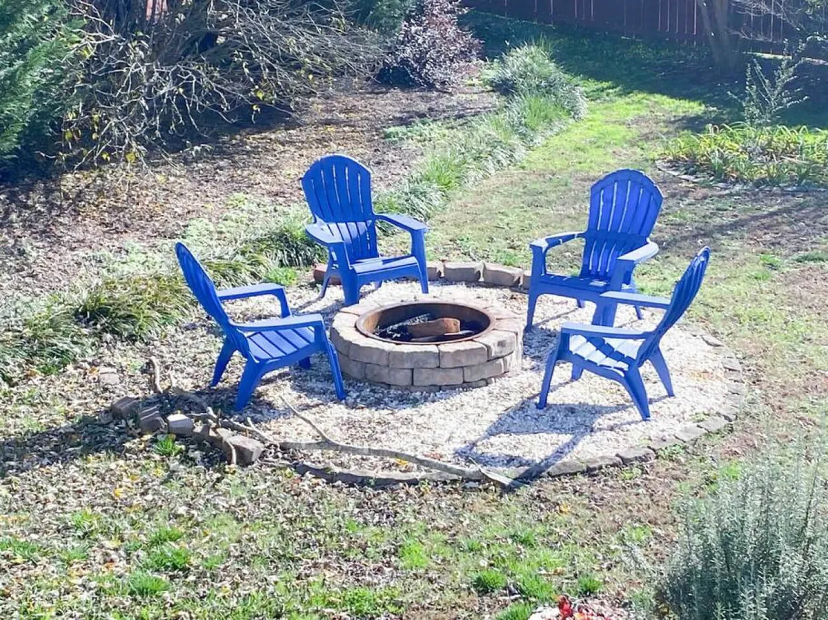 This image features a cozy backyard scene with blue Adirondack chairs arranged around a stone fire pit set within a gravel area. The backdrop includes mature trees and soft lighting, evoking the feeling of evening gatherings. The area is clearly designed for socializing, storytelling, and marshmallow roasting, with ample space for friends and family to gather.