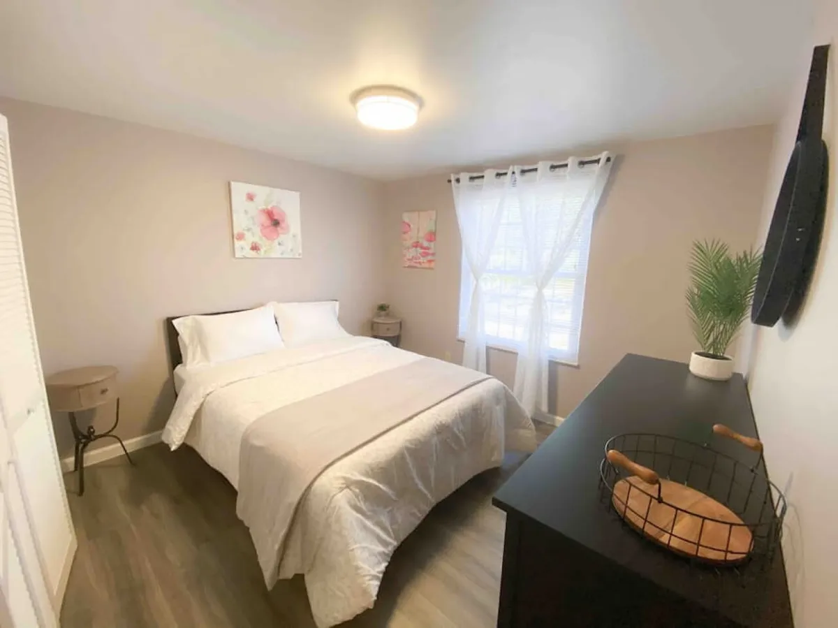A serene bedroom featuring a bed with white bedding, a window with sheer curtains letting in gentle natural light, floral wall art, and a black dresser. The space feels peaceful and inviting, ideal for relaxation.