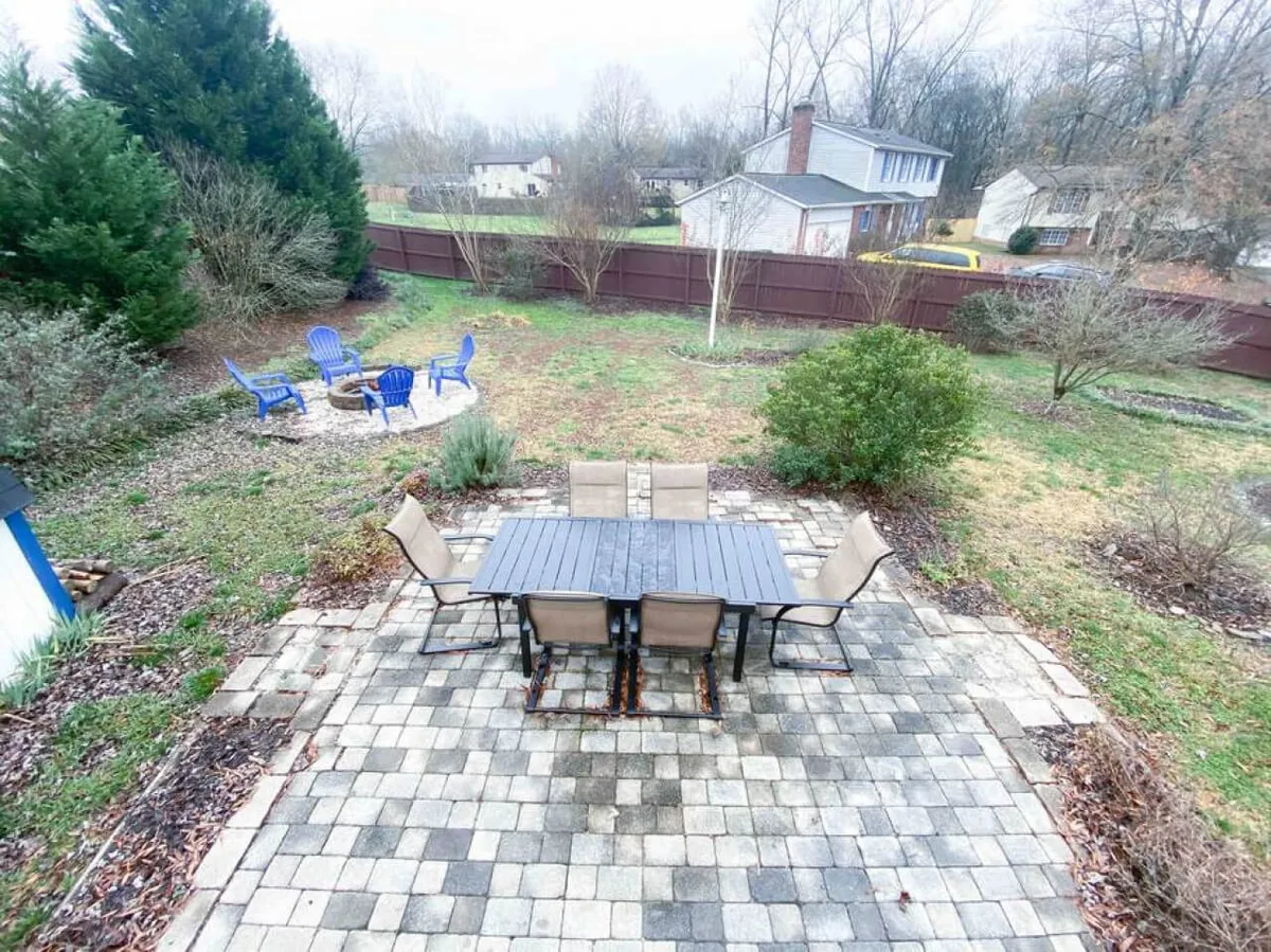 This exterior image features a tranquil backyard setting with a patio table and chairs, blue Adirondack chairs, lush greenery, and a fire pit area. The scene is inviting and highlights outdoor living and relaxation.