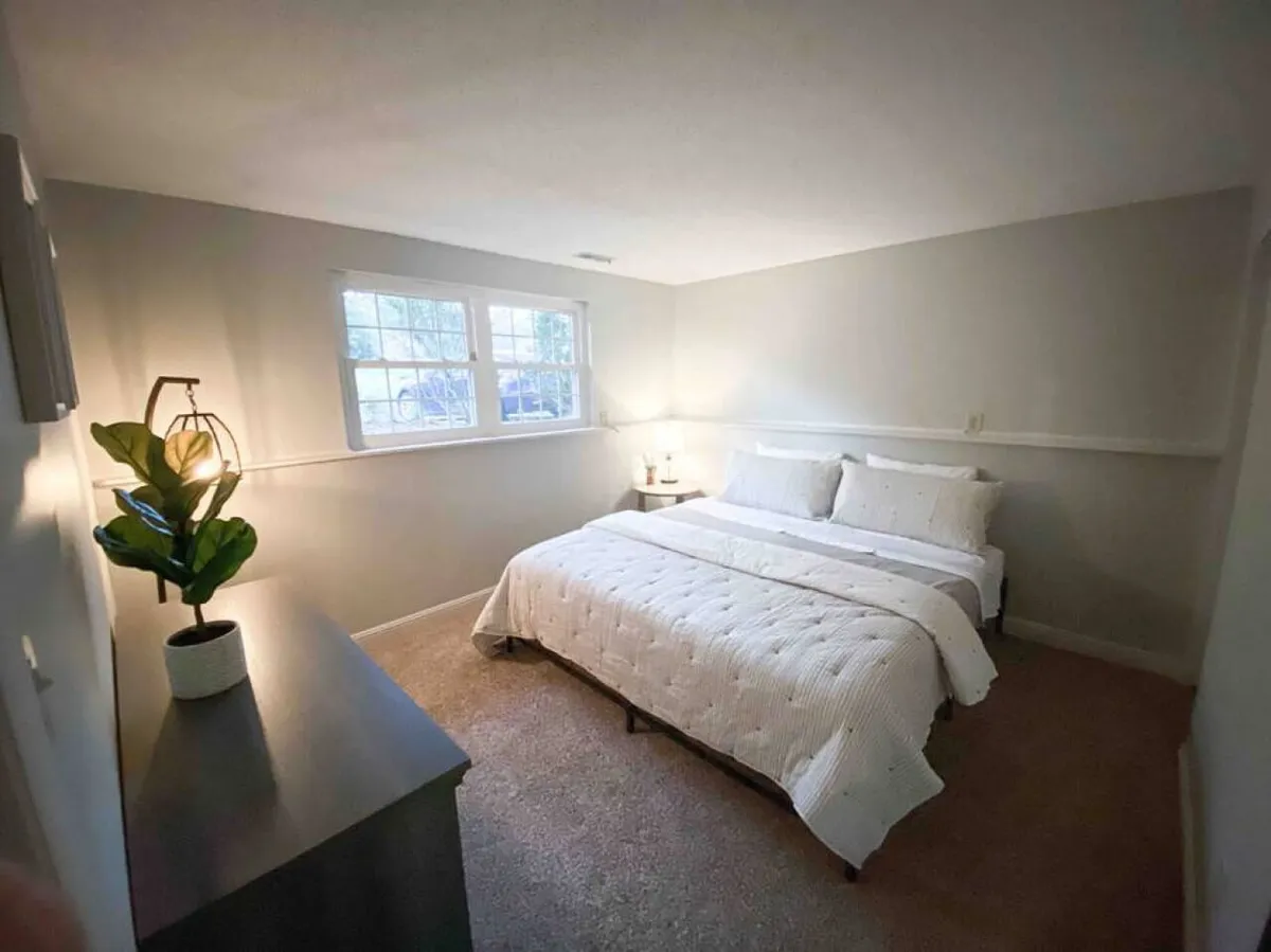 A bright bedroom with a generous bed, large windows that allow sunshine to pour in, a nightstand with a warm-lit lamp, and a touch of greenery from a potted plant. The room is styled for comfort and rejuvenation.