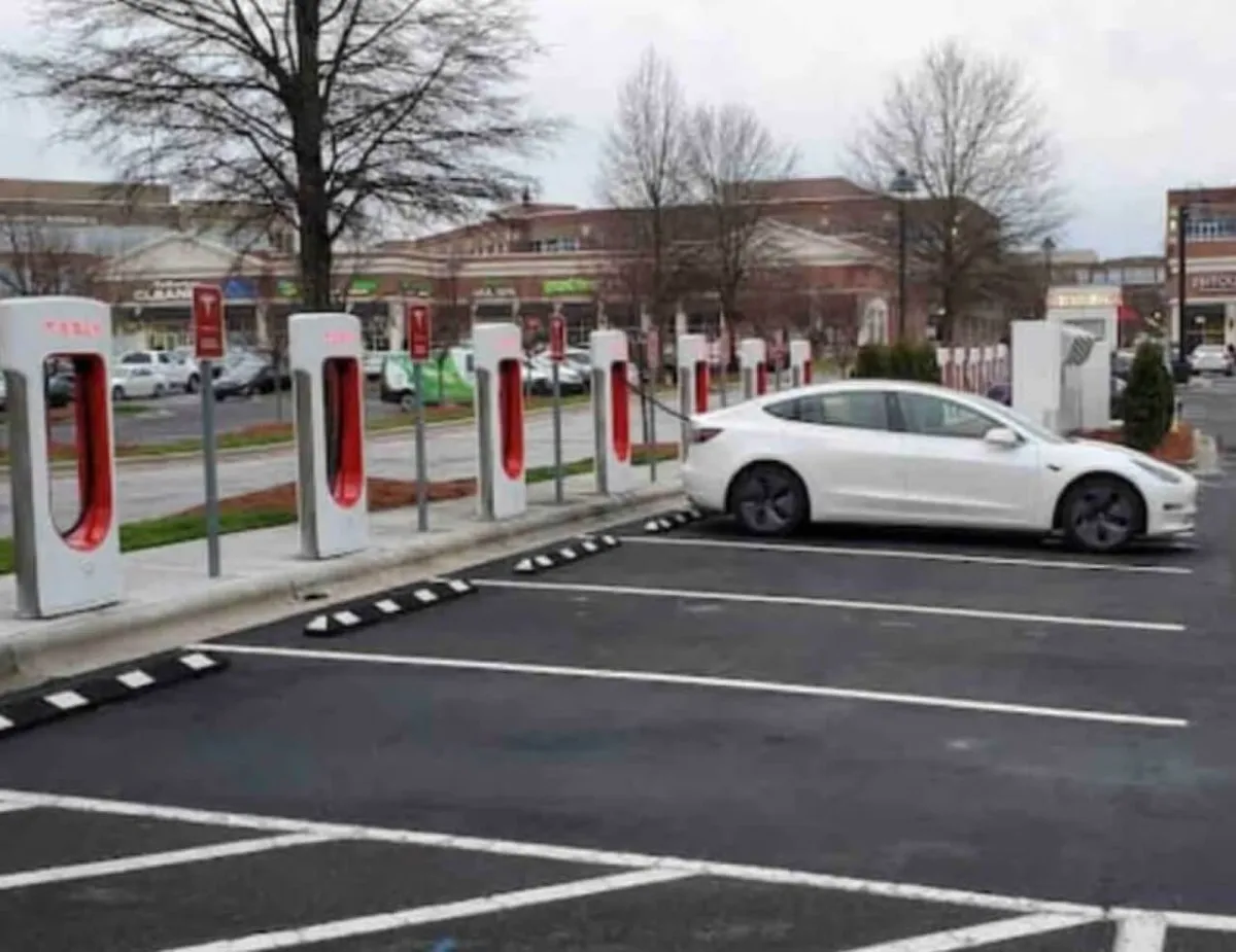 An exterior, urban scene showing several Tesla Superchargers with a white Tesla vehicle parked in front, surrounded by a retail environment. Pedestrians and shoppers can be seen nearby, suggesting a convenient and lively neighborhood.